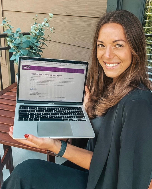 Inspira TLB Founder outside wearing a black blazer, dress pants, sitting down, holding her laptop facing the camera. She is smiling showing her screen their donation to Lupus Foundation of America.